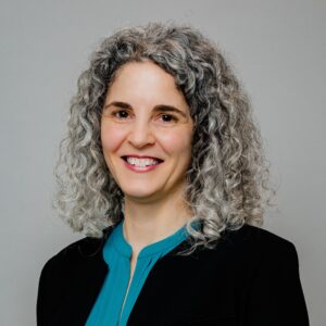 Headshot of fractional general counsel Alisha Bloom, a white woman with dark eyes and salt and pepper shoulder length curly hair, smiling at the camera. Alisha is wearing a dark suit jacket and a teal green blouse.